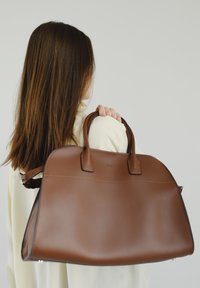 Bruine leren handtas met een gladde textuur, ronde vorm, korte handvatten en een afneembare schouderriem. Kenmerkt subtiele branding en strakke lijnen.