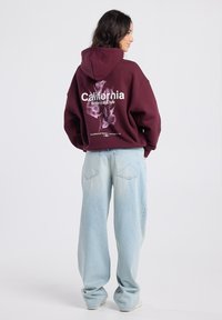 Woman facing away wears maroon hoodie with "California Botanical Club" floral print and light blue wide-leg jeans on white background.