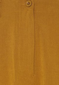 Detaillierte Nahaufnahme eines braunen Stoffes, der einen Knopf, eine Tasche und sichtbare Nähte zeigt. Glatte Textur mit matter Oberfläche.
