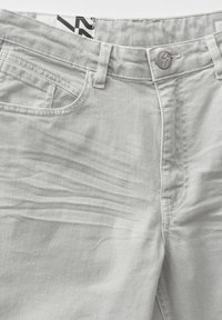 Jean en denim gris clair à l’avant avec fermeture par bouton et passants de ceinture visibles, vue partielle de la ceinture et des poches avant.