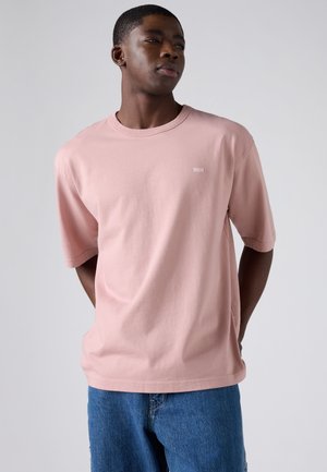 Νεαρός άνδρας που φοράει ένα ανοιχτό ροζ υπερμέγεθος μπλουζάκι και μπλε τζιν, στέκεται με τα χέρια πίσω από την πλάτη, κοιτώντας προς τα πλάγια μπροστά σε απλό φόντο.