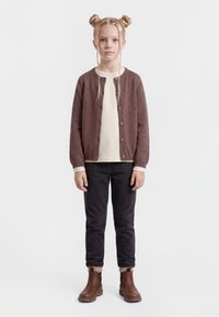 Persona joven con cabello rubio recogido en moños espaciales y trenzas, vistiendo un cárdigan marrón abotonado, camisa color crema, pantalones negros y botas marrones, de pie.