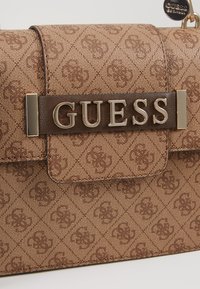 Ruskea käsilaukku, jossa on teksturoitu kuvio, kullanvärinen "GUESS"-logo ja tummanruskea nahkainen yksityiskohta. Magneettinen suljenta ja jämäkkä muotoilu.