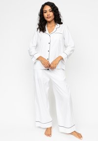 Witte pyjama-set met een knoopsluiting aan de bovenkant, een borstzak, zwarte piping, lange mouwen en ruimvallende broek. Lichte stof.