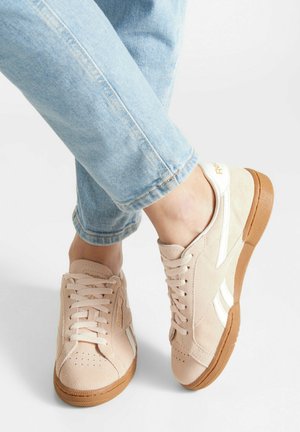 Hellbeige Wildleder-Sneaker mit weißen Streifen und braunen Sohlen, getragen zu hellblauen Jeans, die Füße überkreuzt vor schlichtem grauem Hintergrund.