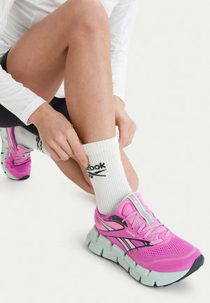 Person adjusting white Reebok sock, wearing bright pink Reebok running shoe, black shorts, and a white long-sleeve top on a white background.