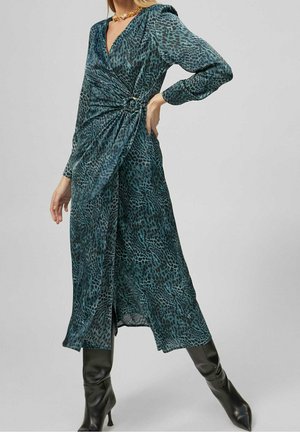 Kobieta w turkusowo-czarnej sukience kopertowej z długimi rękawami i w czarnych wysokich kozakach na obcasie, na jednolitym tle.