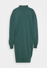 Zelené šaty s vysokým límcem, dlouhými rukávy a volným střihem. Mají žebrované manžety a lem pro zvýšení textury.