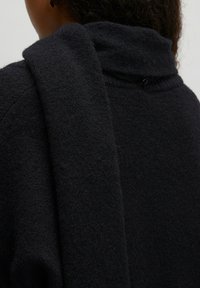 Donker gebreid sweater met een hoge kraag en een langwerpige sjaal die casual is neergelegd. Zachte textuur, subtiele stikdetails en knoopaccent.
