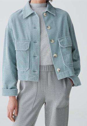Vrouw die een lichtblauwe corduroy jas met grote knopen draagt over een grijze top en grijze broek met zakken.