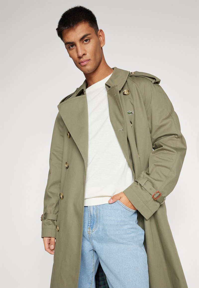 Olivengrüner Trenchcoat mit großen Knöpfen und Schulterklappen, kombiniert mit einem weißen gerippten Pullover und hellblauen Jeans.