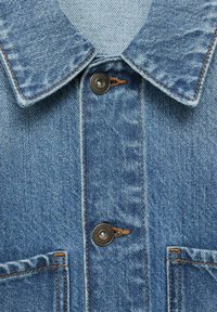 Jachetă din denim de culoare bleu deschis, cu o textură pronunțată, guler, două buzunare frontale și detalii de cusătură maro pe nasturi negri.