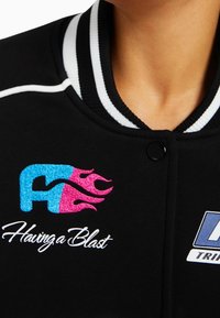 Zwarte varsityjack met witte gestreepte kraag. Bevat een blauw en roze geborduurd logo met vlammen en de tekst "Having a Blast."