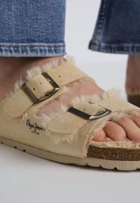 Bež sandale s krznom i dva podesiva remena te metalnim kopčama. Teksturalni, mekani materijal na gornjem dijelu i pluta kao potplat. Logotip brenda vidljiv.