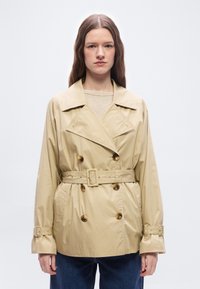 Γυναίκα με μακριά καστανά μαλλιά, φορώντας μπεζ διπλοκουμπωτό παλτό με ζώνη και μπλε τζιν, στέκεται μπροστά σε απλό λευκό φόντο.