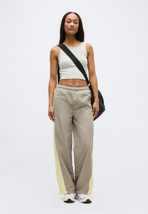 Kobieta stojąca, ubrana w jasnozielony, bezrękawny crop top, beżowe spodnie dresowe z żółtymi paskami, białe trampki oraz czarną torbę na ramię.