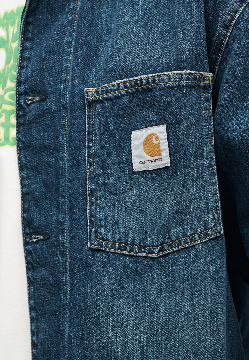 Spijkerjasje zak met beige Carhartt-logo patch en zichtbare stiksels, gedragen over een wit overhemd met groene tekst.