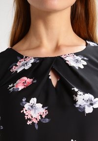 Svart blommig blus med rosa och vita blommor, en nyckelhalsringning och mjukt, slätt tyg med en lätt lyster.