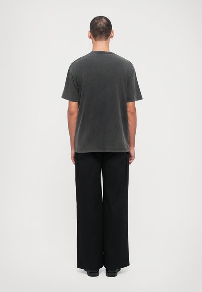 T-shirt cinza escuro de manga curta com um corte descontraído, tecido texturizado, combinado com calças largas pretas e sapatos pretos. Vista a partir de trás.