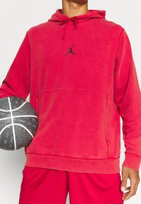 Sweat à capuche en polaire rouge avec poche kangourou, capuche à cordon ajustable et logo noir. Tenant un ballon de basket noir à surface texturée.