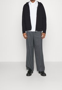 Felpa nera con zip sopra una maglietta polo bianca con un logo, abbinata a pantaloni larghi grigi e scarpe nere, su uno sfondo bianco.