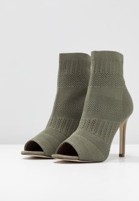 Bottines ouvertes vertes à talon aiguille avec un dessus en tricot texturé, présentent un talon haut et des rayures horizontales.