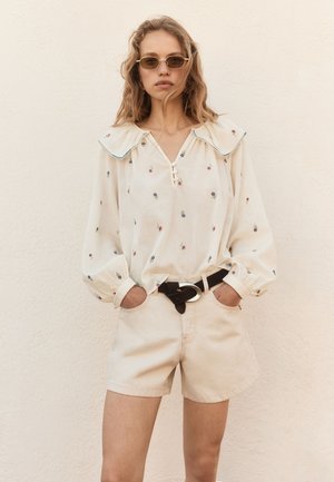 Ung kvinde med bølget hår iført solbriller, en hvid bluse med blomstermønster og stor krave, beige shorts og et sort bælte står op ad en væg.