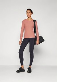 Ροζ μπλούζα με μακριά μανίκια και υφή σχεδίου, σε συνδυασμό με μαύρο κολάν και μαύρα αθλητικά παπούτσια. Το μοντέλο κρατά μια μαύρη τσάντα γυμναστηρίου.