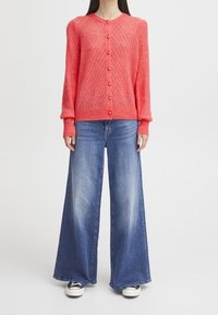 Cardigan lavorato a maglia corallo con motivo testurizzato e scollatura rotonda, chiuso con bottoni davanti. Abbinato a jeans a gamba larga blu chiaro e sneaker bianche.