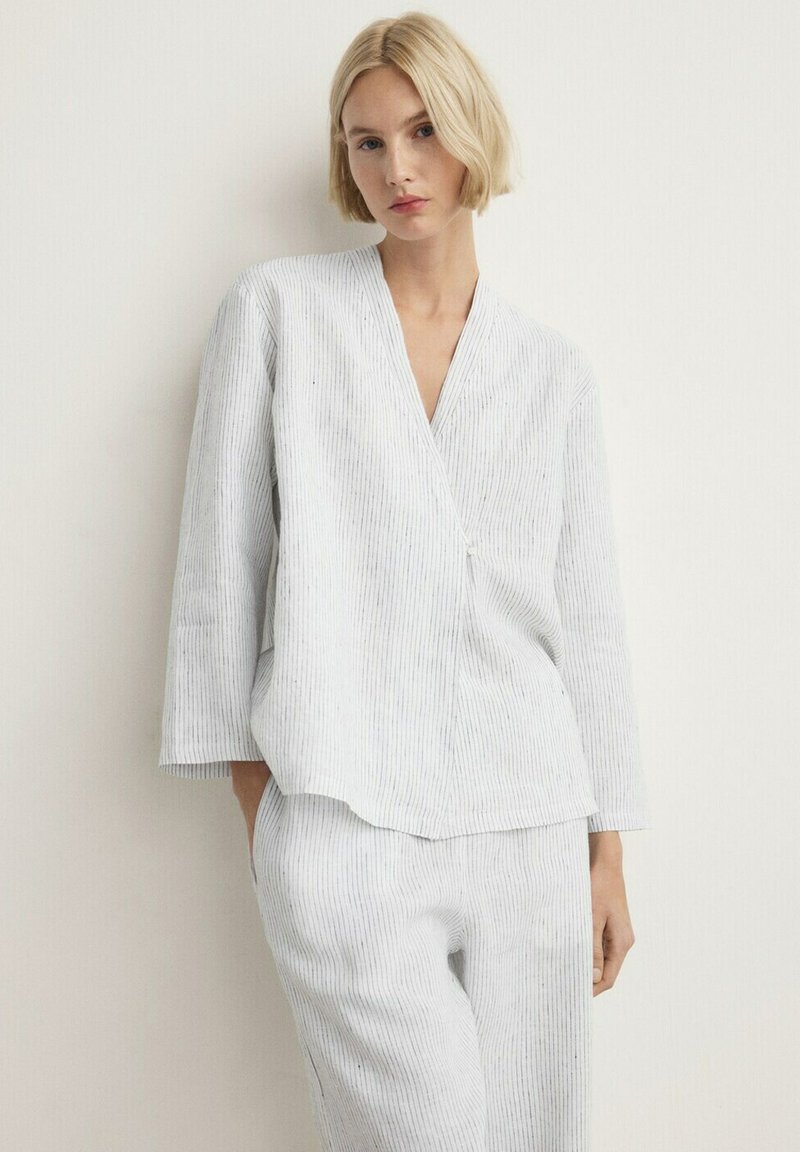 Blondínka oblečená v svetlosivej, prúžkovanej, dlhorukávej zavinovacej blúzke a voľných nohaviciach v rovnakej farbe, stojaca pred jednoduchou bielou stenou.