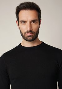 Mężczyzna z krótkimi ciemnymi włosami i brodą, ubrany w czarny sweter z okrągłym dekoltem, patrzący prosto w obiektyw na tle jednolitego jasnego tła.