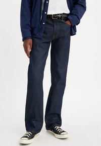 Jean en denim bleu foncé avec une coupe droite, présentant des coutures orange, une fermeture éclair et porté avec des baskets en toile noires.