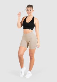 Czarny sportowy biustonosz i beżowe szorty z wysokim stanem. Tkanina wygląda na gładką i elastyczną. Modelka ma na sobie białe buty sportowe.