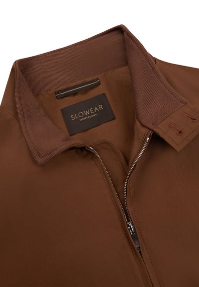 Giacca marrone con collo a coste, cerniera metallica parzialmente aperta e etichetta nera con scritta dorata "SLOWEAR MONTEDORO" all'interno del collo.