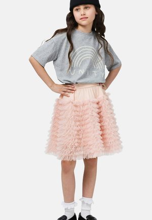 Dziewczyna w czarnej czapce typu beanie, szarej koszulce z napisem "UNITY", różowej falbankowej spódnicy, białych falbaniastych skarpetkach i czarnych butach, stojąca z rękami na biodrach.