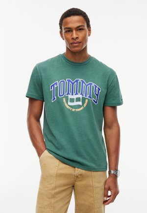 Άνδρας με πράσινη μπλούζα Tommy Jeans και μπεζ παντελόνι με το ένα χέρι στην τσέπη σε λευκό φόντο