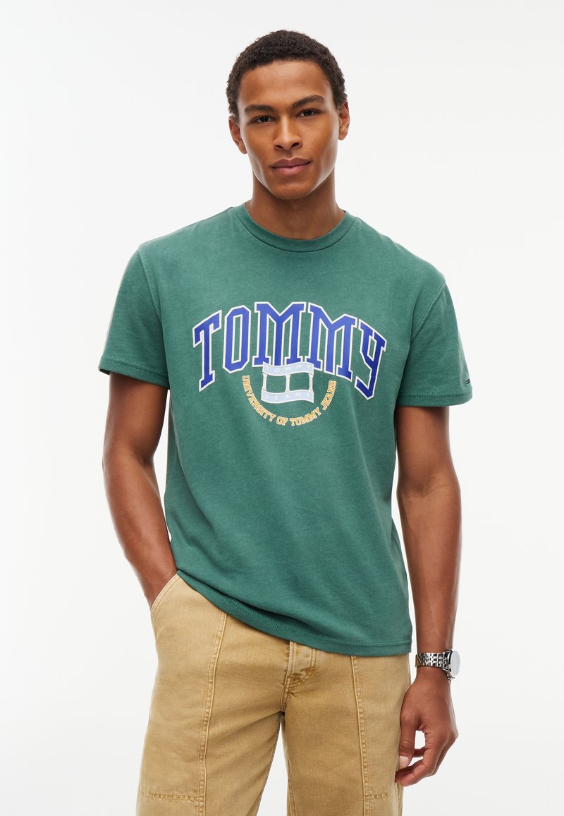 Άνδρας με πράσινη μπλούζα Tommy Jeans και μπεζ παντελόνι με το ένα χέρι στην τσέπη σε λευκό φόντο