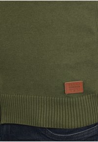 Olivegrüner Strickpullover mit geripptem Saum, glatter Textur und einem rechteckigen braunen Webetikett mit Markennamen und Gründungsjahr.