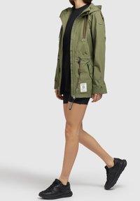 Donna che cammina indossando una giacca con cappuccio verde oliva, pantaloni corti neri, una maglietta nera lunga e sneakers nere su uno sfondo grigio chiaro.