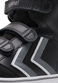 Schwarze synthetische Sneaker mit grauen Akzenten, zwei Klettverschlüssen und auffälligen weißen Zickzackmustern an der Seite.