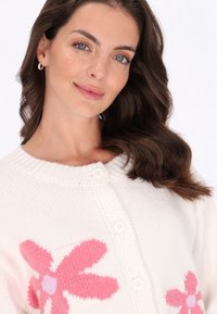 Bílý pletený cardigan se žebrovaným výstřihem, velkým růžovým květinovým vzorem a předním zapínáním na knoflíky. Hladká struktura, útulný design.