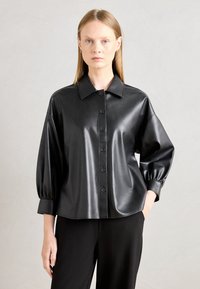 Zwarte faux leren shirt met een kraag, knoopsluiting aan de voorkant en losse, gepofte mouwen; gladde textuur en glanzende afwerking.