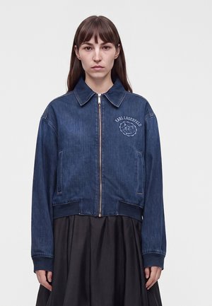 Kvinde iført en blå denim jakke med lynlås med en lille patch og tekst på venstre bryst, kombineret med et sort skørt.