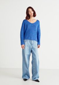 Blå strikket genser med v-hals, parret med lyseblå, vide jeans og svarte spisse sko. Teksturisert stoff, løs passform.