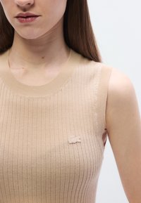 Top sin mangas beige con textura acanalada y cuello redondo, con un pequeño logo bordado en el lado izquierdo. Textura suave y diseño ajustado.