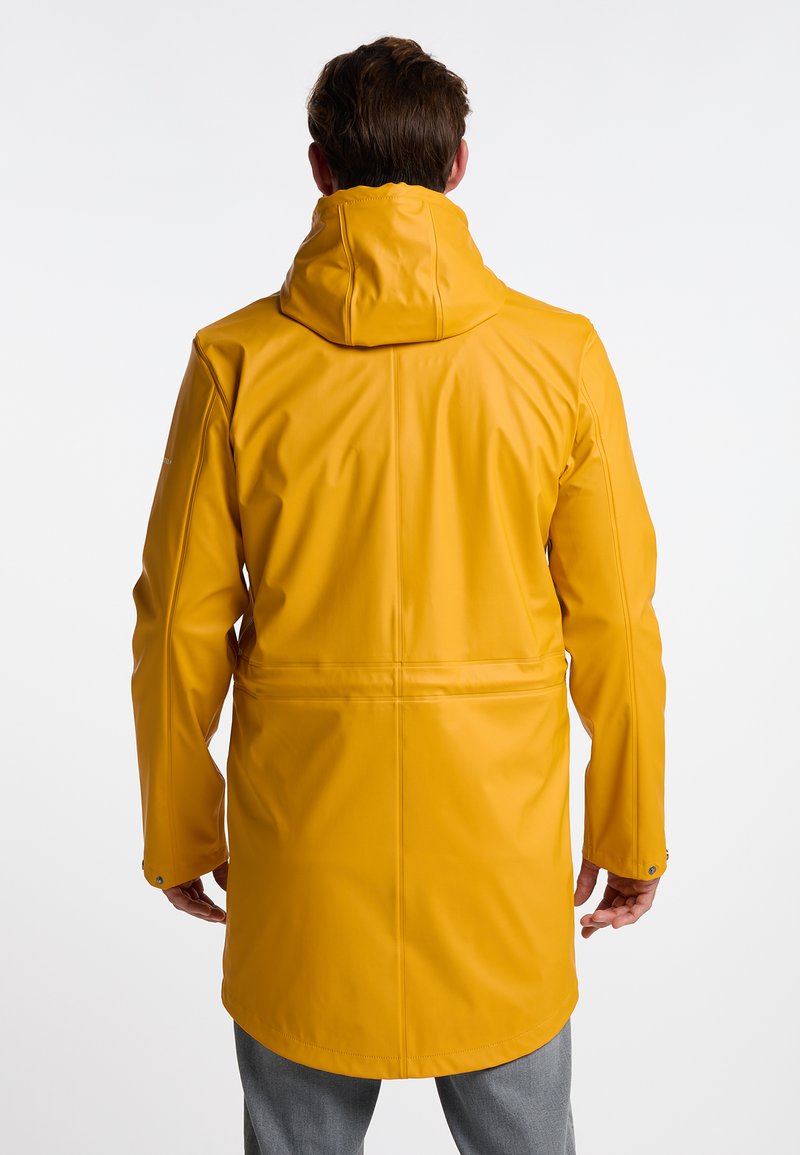 Man met een lange gele regenjas met een capuchon, staand met zijn rug naar de camera tegen een effen witte achtergrond.