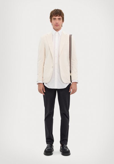 Light beige blazer over a white button-up shirt, black trousers, and glossy black shoes. Features a shoulder bag with a patterned strap.