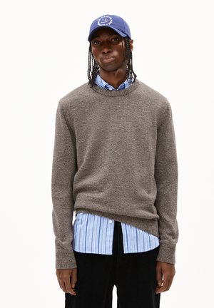 Pull en tricot marron texturé, superposé à une chemise rayée bleu clair. Casquette bleue avec un design de smiley, pantalon en velours côtelé noir porté.