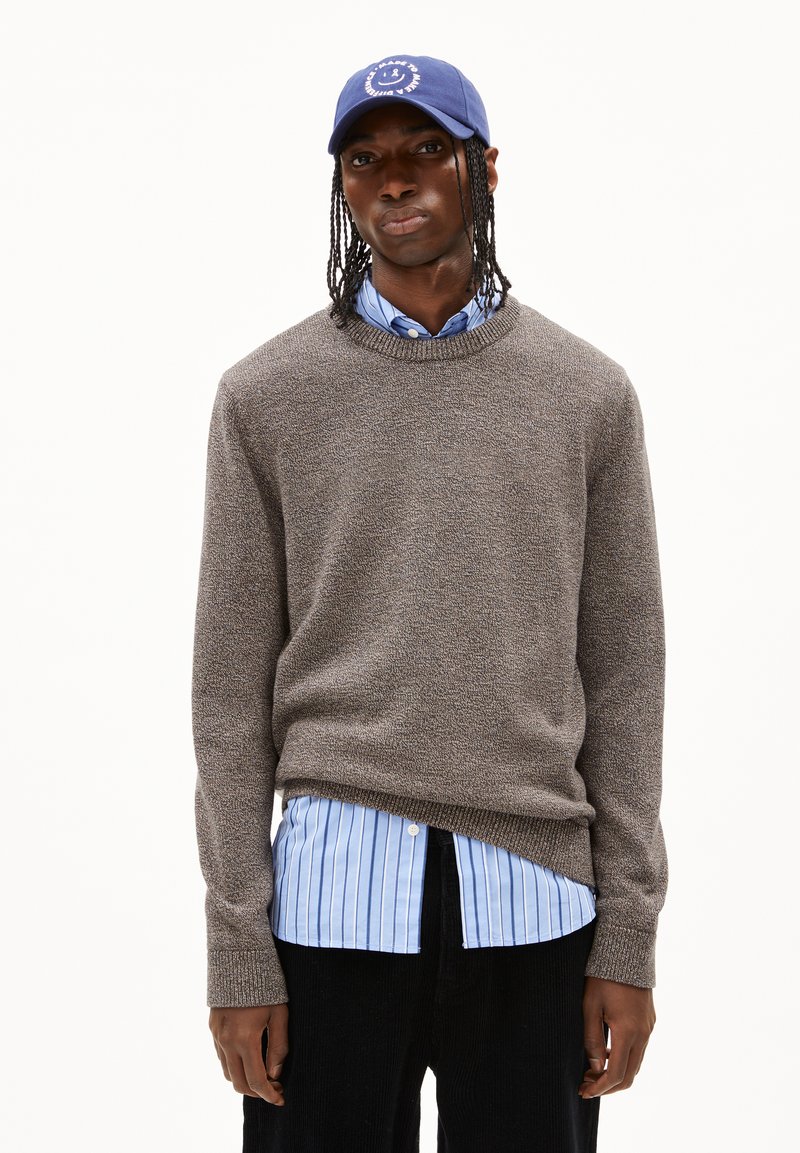 Pullover aus strukturiertem braunem Strick, über einem hellblau gestreiften Hemd getragen. Blaue Mütze mit einem Smiley-Motiv, schwarze Lodenhose wird getragen.