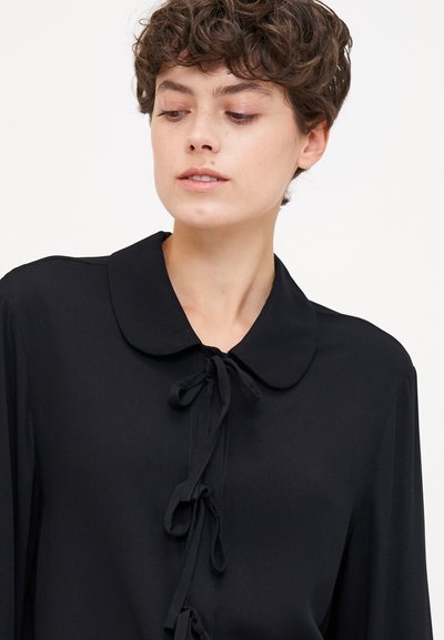 Mustmustelne blouse, millel on kõver kaelus, esitõmblused ja pikad varrukad. Valmistatud siledast kangast, millel on matt viimistlus.